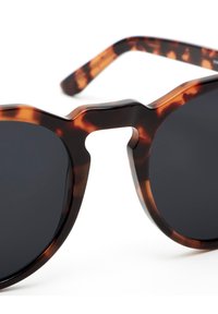 Tortie stila saulesbrilles ar apaļām melnām lēcām, ar biezu, matētu rāmi un izteiktu tiltu; eleganti, noapaļoti tempļi.