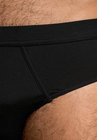 Slip da bagno neri con una superficie liscia, caratterizzati da un ampio elastico in vita e cuciture a contrasto. Ingrandimento del lato con peli corporei visibili.