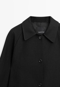 Zwarte jas met een reverskraag, voorzien van een knopenfront en een gestreepte voering. Het materiaal lijkt gestructureerd en glad. Merketiket zichtbaar.