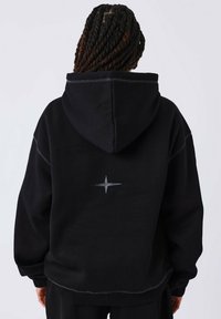 Una persona con capelli intrecciati indossa una felpa nera con un logo riflettente a forma di stella centrato sulla schiena, in piedi contro uno sfondo semplice.