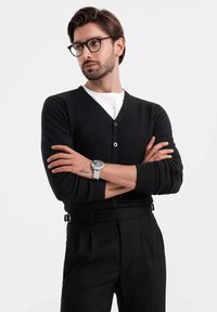 Svart cardigan med V-ringning, figurnära passform och knappstängning, lagd över en vit T-shirt. Modellen bär svarta byxor och en silverfärgad klocka.