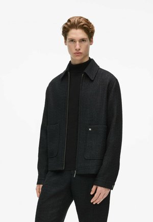 Young man with light brown hair wears a textured dark jacket with large front pockets over a black turtleneck and matching dark pants.