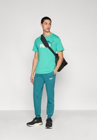 Τιρκουάζ μπλουζάκι με λευκό λογότυπο "PUMA", σε συνδυασμό με πράσινο-μπλε παντελόνι φόρμας με το λογότυπο. Φορεμένα με μαύρη τσάντα ώμου και μαύρα αθλητικά παπούτσια.