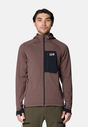 Mann trägt eine braune Jacke mit Reißverschluss, schwarzer Brusttasche und Daumenlöchern an den Ärmeln vor einem schlichten Hintergrund.
