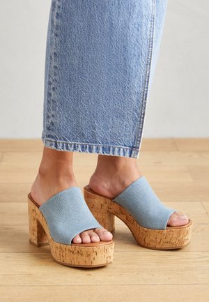 Pieds portant des sandales à plateforme à bout ouvert avec talons en liège et brides en tissu bleu clair, associées à un jean court en denim bleu clair.