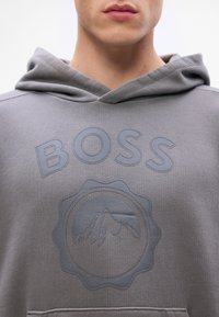 Hanorac gri texturat cu un logo în relief care include numele brandului "BOSS" și un design de munte într-un cerc.