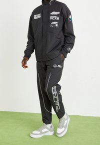 Svart vindjacka med flera logotyper och vita accenter, tillsammans med matchande svarta byxor med vita kantlinjer och en logotyp, bärs med grå sneakers.