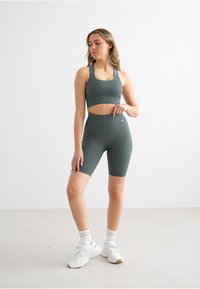 Donkergroen geribbelde sportbeha en bijpassende fietsbroek met hoge taille. Model draagt witte sneakers en sokken met gouden strepen.