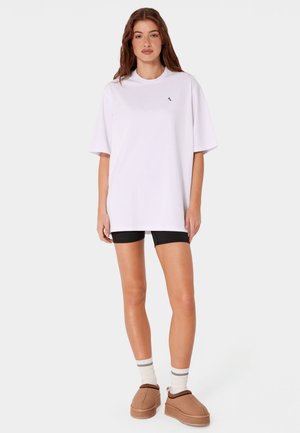 Młoda kobieta stoi ubrana w oversize'ową białą koszulkę, czarne spodenki rowerowe, białe skarpetki i beżowe wsuwane buty na jednolitym tle.