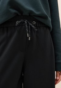 Schwarze Trainingshose mit elastischem Bund, ausgestattet mit einem Kordelzug mit metallischen Enden und einer glatten Textur. Dunkelgrünes Oberteil teilweise sichtbar.