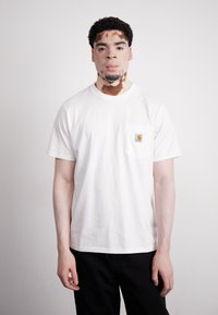 Valkoinen puuvillapaita, jossa on pyöreä pääntie ja tasku, jossa on neliömäinen Carhartt-logo. Paitaa käytetään mustien housujen kanssa, ja henkilö seisoo yksinkertaista taustaa vasten.