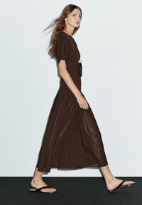 Brun maxi klänning med en figurnära topp och en flödande, lager-på-lager kjol. Modellen bär svarta flata sandaler och ett långt hänge. Slät textur.