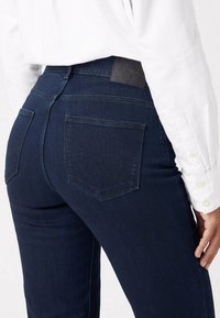 Donkerblauwe spijkerbroek van denim met achterzakken en een zwart leren taillebandlabel. De stof lijkt glad met een lichte rek.