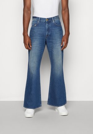 Personne porte un jean flare bleu avec des détails délavés, des baskets blanches et un haut blanc, debout devant un fond clair uni.