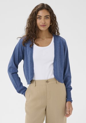 Jauna moteris su garbanotais rudais plaukais, dėvinti mėlyną trumpą kardiganą, baltą palaidinę ir smėlio spalvos aukštakulnes kelnes, stovinti viena ranka kišenėje.