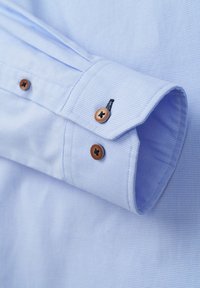 Himmelblaues karierter Hemdarm mit zwei braunen Knöpfen und schwarzen Nähdetails. Glatte Stoffstruktur, taillierte Ausführung.