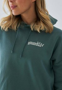 Ciemnozielona bluza z kapturem, wykonana z miękkiego materiału, z białym logo "królewna pilates" oraz regulowanymi sznurkami przy kapturze.