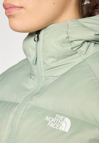 Lichtgroene pufferjas met hoge kraag, voorrits en logo op de linker borst. Vlotte textuur met gestikte panelen.