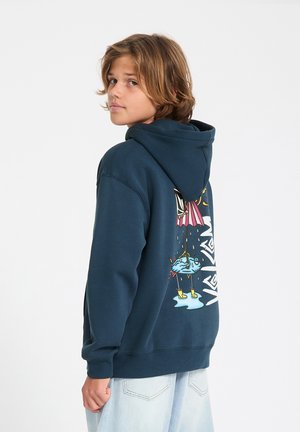 Tienerjongen in een navy hoodie met een kleurrijke vis- en parapluafbeelding op de rug, kijkt over zijn schouder tegen een effen achtergrond.