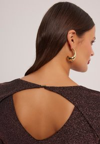 Ciemnofioletowa sukienka o fakturze z otwartym plecami, z zapięciem przy dekolcie i dużymi złotymi kolczykami w kształcie kół.