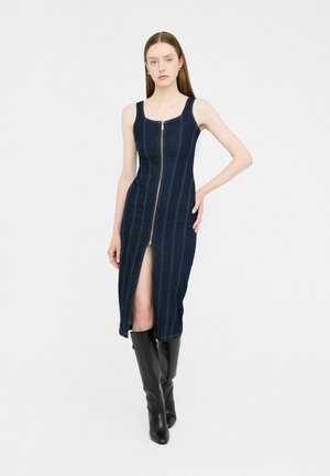 Das Model posiert in einem figurbetonten, dunklen Jeanskleid mit einem Reißverschluss vorne und einem seitlichen Schlitz, kombiniert mit kniehohen schwarzen Stiefeln. Der weiße Hintergrund hebt den Fokus hervor.