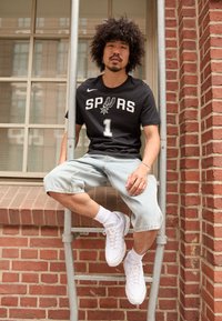 Sort sports skjorte med "SPURS" og nummer "1" trykt i hvid, parret med lyse denimshorts og hvide sneakers med ankelstrømper.