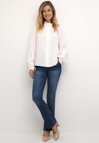 White blouse with a ruffled front, long sleeves, and high neckline, paired with dark blue jeans and nude high-heeled shoes.