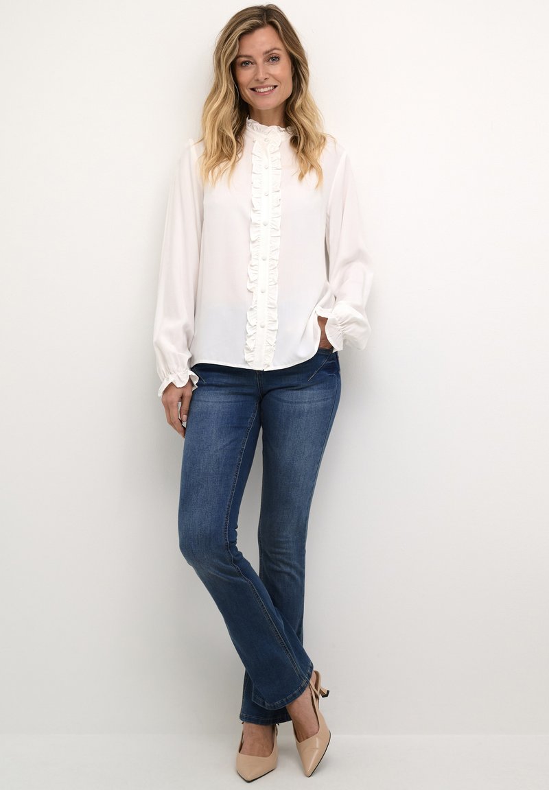 White blouse with a ruffled front, long sleeves, and high neckline, paired with dark blue jeans and nude high-heeled shoes.