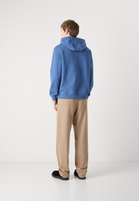 Blå hettegenser med stikkede mansjetter, sammen med løse beige bukser og svarte sko, sett bakfra mot en nøytral bakgrunn.