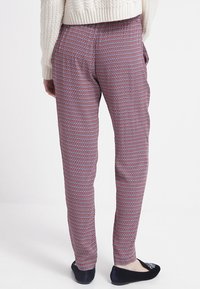 Persoon in slim-fit broek met een klein geometrisch patroon in rood, wit en blauw, gecombineerd met een crèmekleurige trui en zwarte ballerina's.