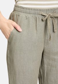 Světlé šedé lněné kalhoty s elastickým pasem, detail s tkaničkou a bočními kapsami. Textura látky se zdá být měkká a mírně texturovaná.