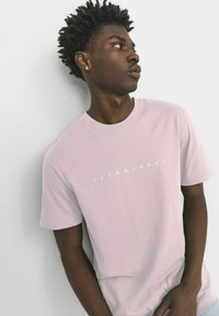 Hellrosa T-Shirt mit kurzen Ärmeln aus Baumwolle, das ein weißes Logo "JACK&JONES" über der Brust zeigt, vor einem neutralen Hintergrund.