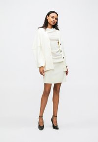 Λευκό oversized cardigan πάνω από ένα υφασμάτινο ανοιχτό πουλόβερ, συνδυασμένο με μια ριχτή φούστα μέχρι το γόνατο στο ίδιο χρώμα και μαύρες γόβες.