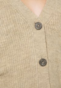 Cárdigan de punto beige con escote en V y un acabado texturizado. Presenta un botón gris en el frontal para el cierre. Diseño simple, material suave.