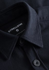 Camicia button-up di colore blu scuro con tessuto testurizzato. Presenta un colletto classico, etichetta con marchio e dettagli dei bottoni sulla patta.