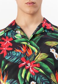 Homme portant une chemise noire à boutons ornée de fleurs tropicales rouges, orange et blanches ainsi que de feuilles vertes. Vue dégagée sur le col et le haut de la poitrine.