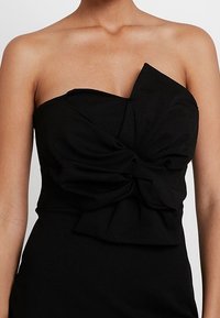 Robe noire sans bretelles avec un grand nœud torsadé sur la poitrine, portée par une personne avec les épaules et les bras supérieurs découverts.