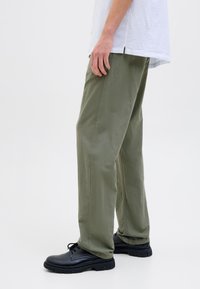 Persona vestida con pantalones holgados color verde oliva, zapatos negros con cordones y una camisa blanca, de pie contra un fondo blanco liso.
