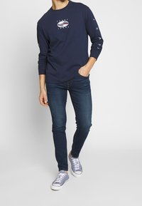 Marinblå långärmad T-shirt med en tryckt logotyp i mitten och grafiska detaljer längs ärmarna, kombinerad med mörkblå slim-fit jeans och sneakers.