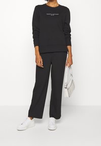 Felpa nera e pantaloni a gamba larga realizzati in un tessuto morbido. Sneakers bianche e una piccola borsa texturizzata completano l'outfit. Logo visibile sulla felpa.