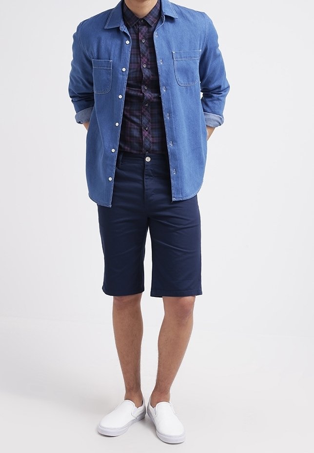 Chemise en jean boutonnée avec manches retroussées portée par-dessus une chemise à carreaux, associée à un short bleu marine et des chaussures blanches sans lacets. Ensemble simple et décontracté.