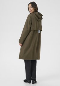 Jachetă impermeabilă de culoare verde măsliniu cu glugă, având un design scurt în spate, mâneci lungi și buzunare laterale, asortată cu blugi negri și pantofi cu toc.