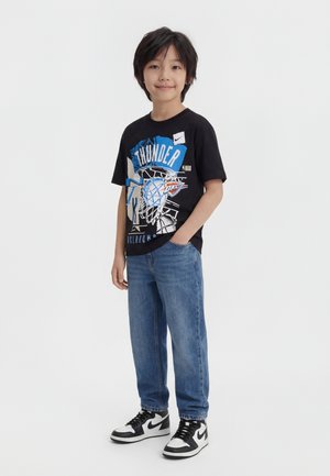 Junge mit schwarzen Haaren, der ein schwarzes Oklahoma City Thunder NBA-Grafik-T-Shirt, blaue Jeans und schwarz-weiße Nike-Sneaker trägt, mit der Hand in der Tasche stehend.