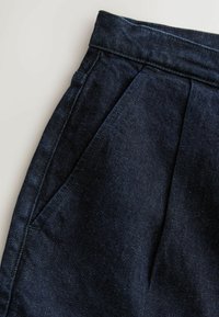 Pantalon en denim bleu foncé avec plis à l'avant et poche latérale sur un fond clair, montrant la texture du tissu et les détails des coutures.