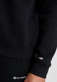 Sudadera negra fabricada con una mezcla de algodón, con puños y dobladillo de canalé, y un pequeño logo rojo y blanco en la muñeca izquierda.