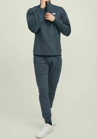 Mörkgrå tröja med hög krage och dragkedja fram, ihopparad med matchande joggingsbyxor. Har ribbade mudd och vita sneakers.