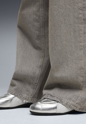 Graue Jeans mit weitem Bein bedecken glänzende silberne Sportschuhe mit schwarzen Sohlen, die ein kleines Logo-Akzent besitzen. Der Stoff hat eine weiche Textur.