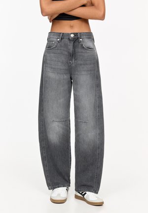 Osoba nosząca luźne, wyblakłe czarne jeansy z wysokim stanem oraz białe trampki z czarnymi paskami, stojąca z założonymi ramionami na tle białym.