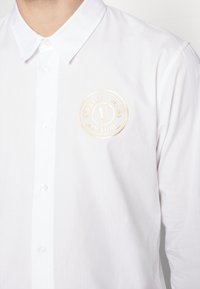 Camicia bianca con bottoni realizzata in un tessuto liscio, caratterizzata da un logo circolare ricamato in oro con la scritta "Versace Jeans Couture".