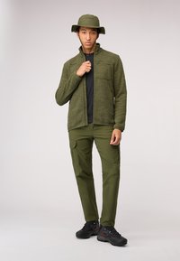 Giacca in pile verde oliva con colletto alto, zip frontale e tasca sul petto. Abbinata a pantaloni cargo coordinati e scarpe da escursionismo nere.